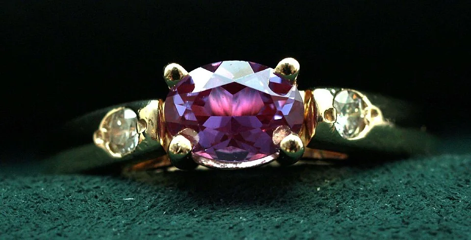 Purple faceted center stone with pale round side stones in a yellow-metal ring.