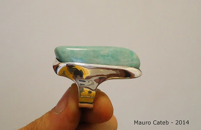 Side view of a pale blue-green stone ring in a curved metal setting, held between fingertips.