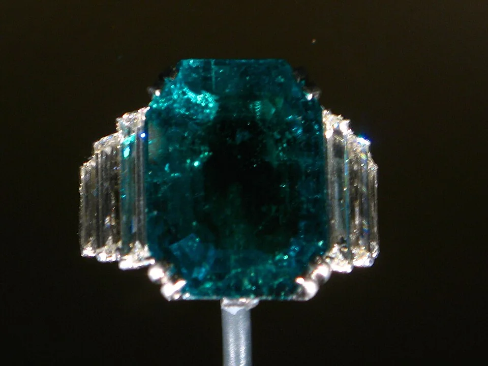 Rectangular deep-green stone with clear side stones in a ring against a dark background.