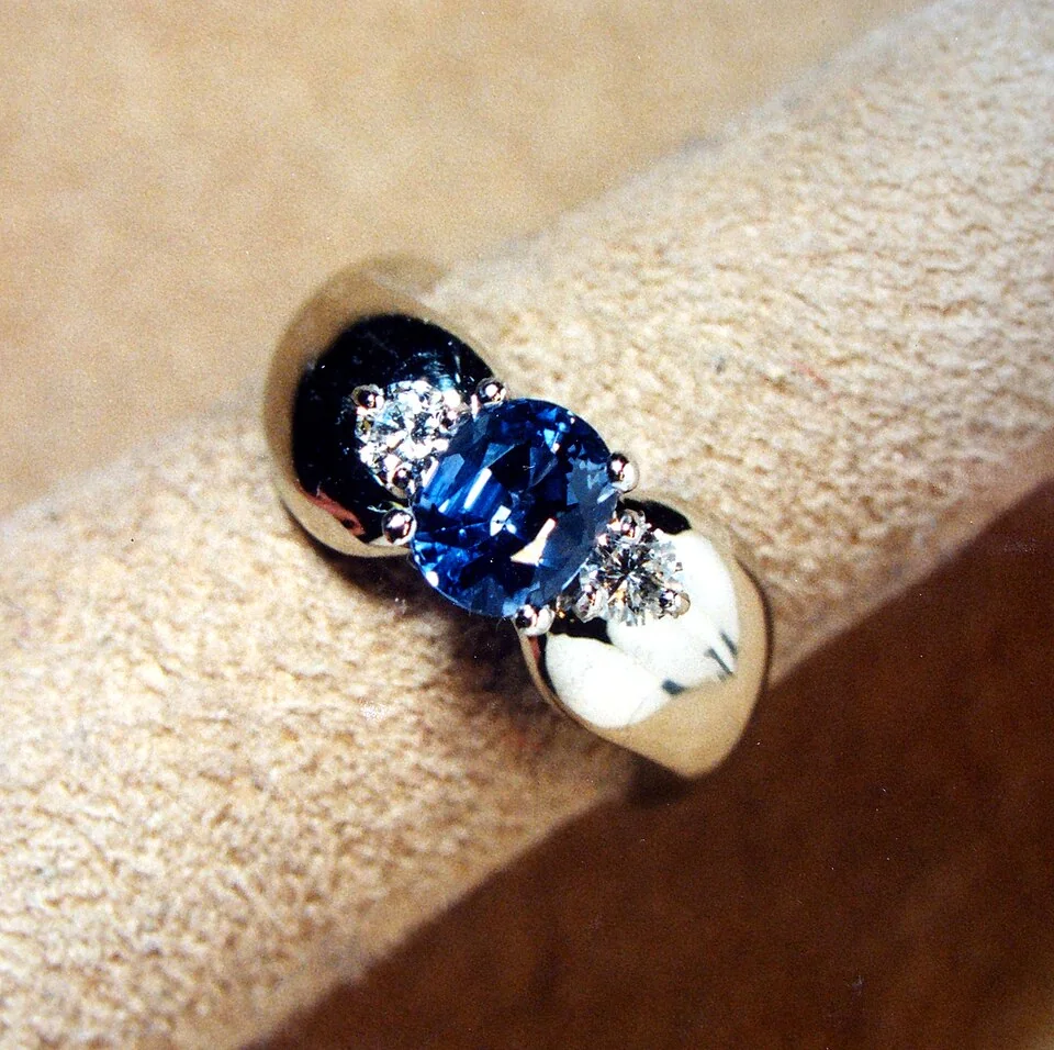 Round blue faceted stone between pale side stones in a broad metal ring.