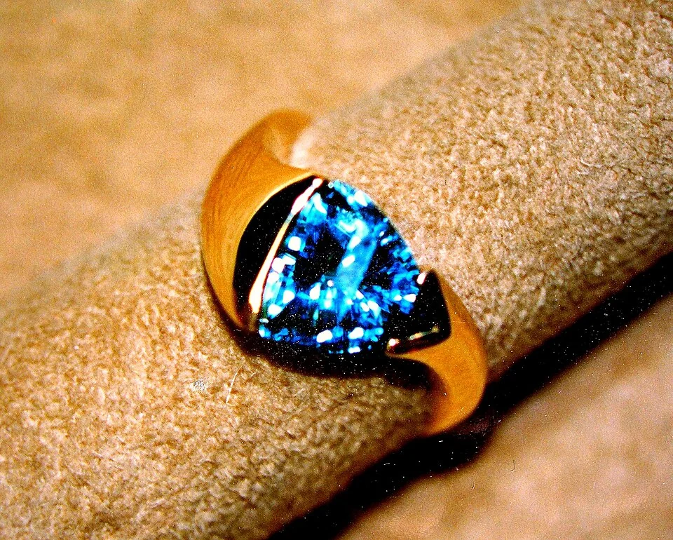 Bright-blue triangular faceted stone in a broad yellow-metal ring.