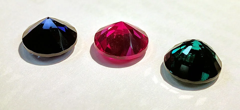 Three loose faceted stones, blue, pink-red, and green, on a white surface.