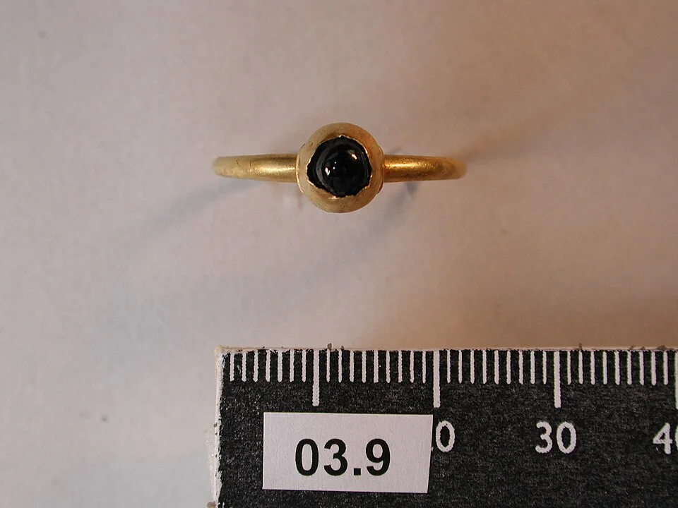 Slender yellow-metal band with a small dark round stone in a simple bezel, beside a ruler.