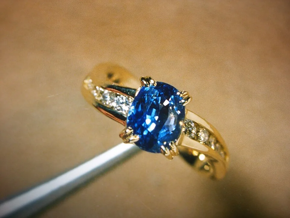 Bright-blue faceted stone with pale side stones in a yellow-metal ring.