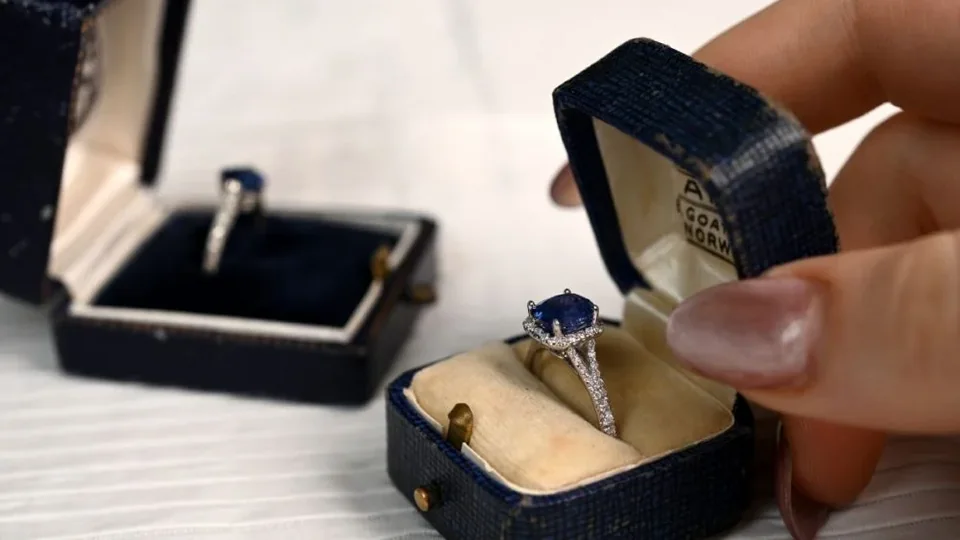Hands opening a ring box containing a blue-stone ring beside a second box.